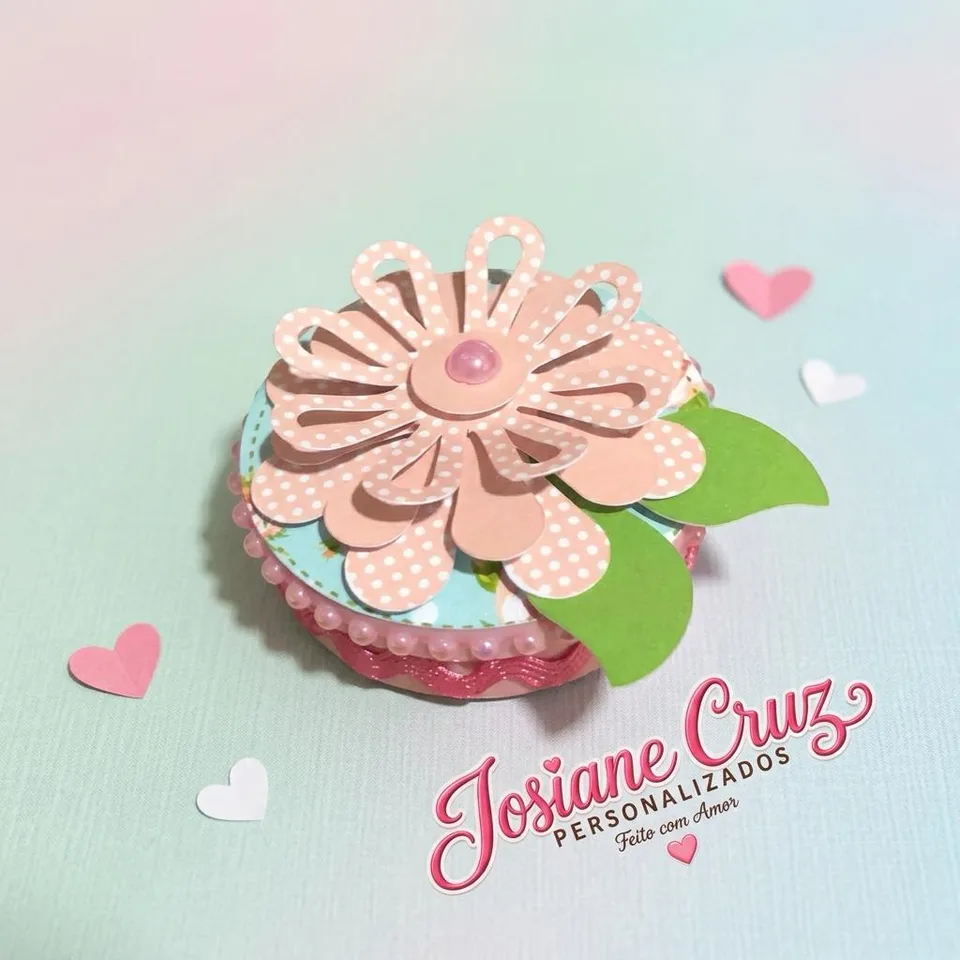 Latinha decorada com flor rosa em camadas e folhas verdes sobre fundo claro.